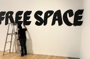 A person paints in black letters "free space" on a white wall