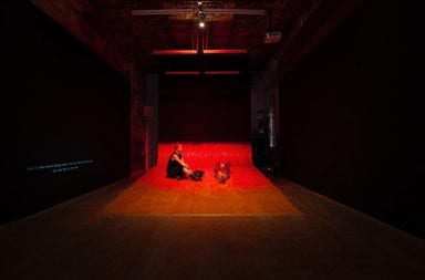 A gallery space is darkened, with black walls, wooden floor, and red light. White open captions are projected dimly onto the left wall. A wooden ramp curls up the back wall. Two people are mid-way up the ramp; the person on the right reclines vertically with arms behind their head, and the person on the right sits upright facing their friend. Two corridors flank the back wall. A soft red glow fills the space behind the back wall