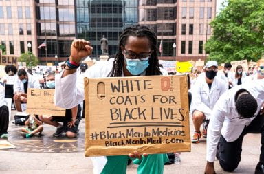 An African American male doctor kneels down with a first raised while holding a sign