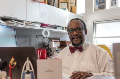 A black man wearing a white shirt and red bow tie smiles for the camera in his office