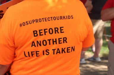back of person wearing orange shirt that reads "#OSUPROTECTOURKIDS BEFORE ANOTHER LIFE IS TAKEN"