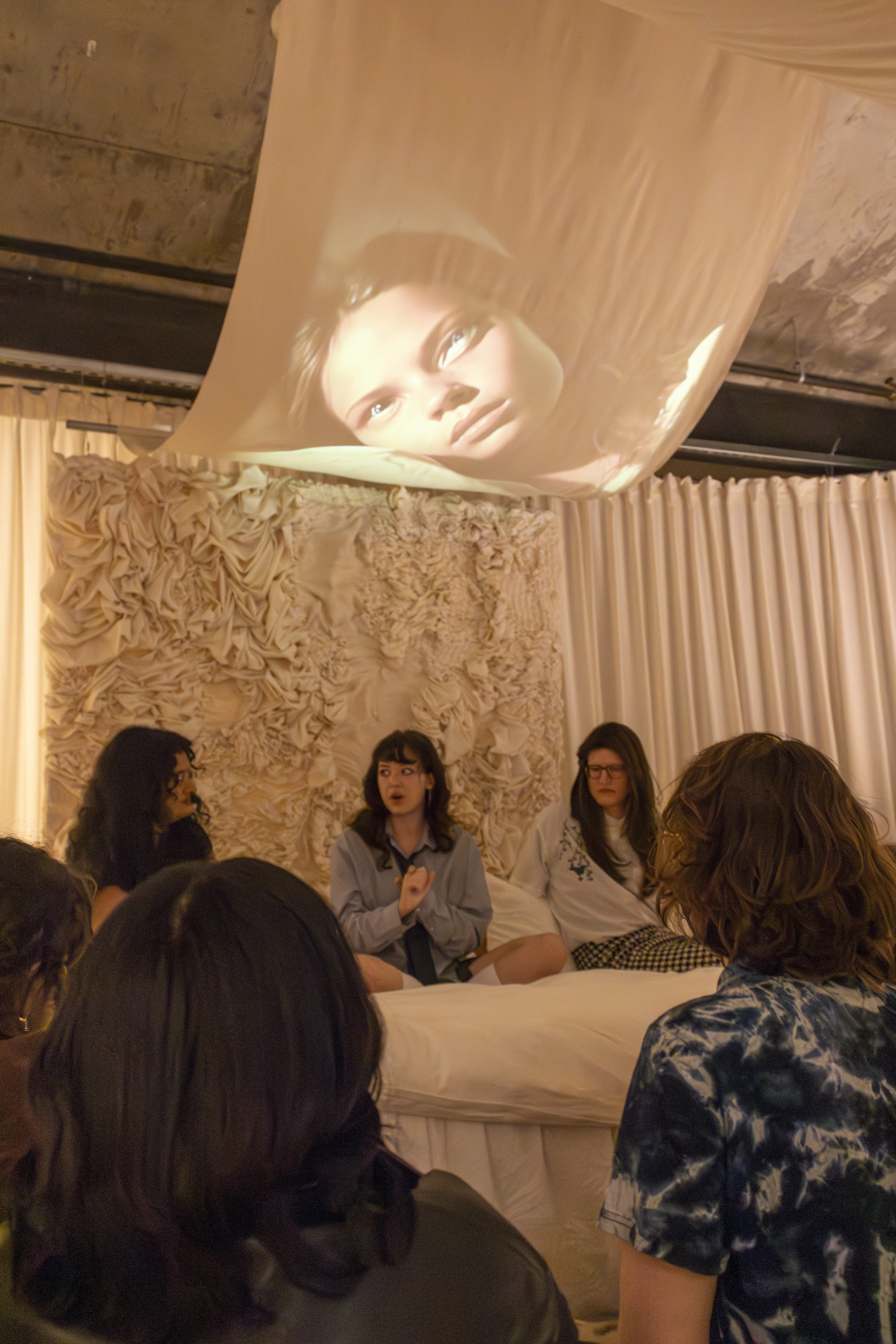 Artist Rayne Fisher-Quann sits on a bed as images of feminine media flash above her on a canopy. Credit: Sandra Fu | Lantern Reporter